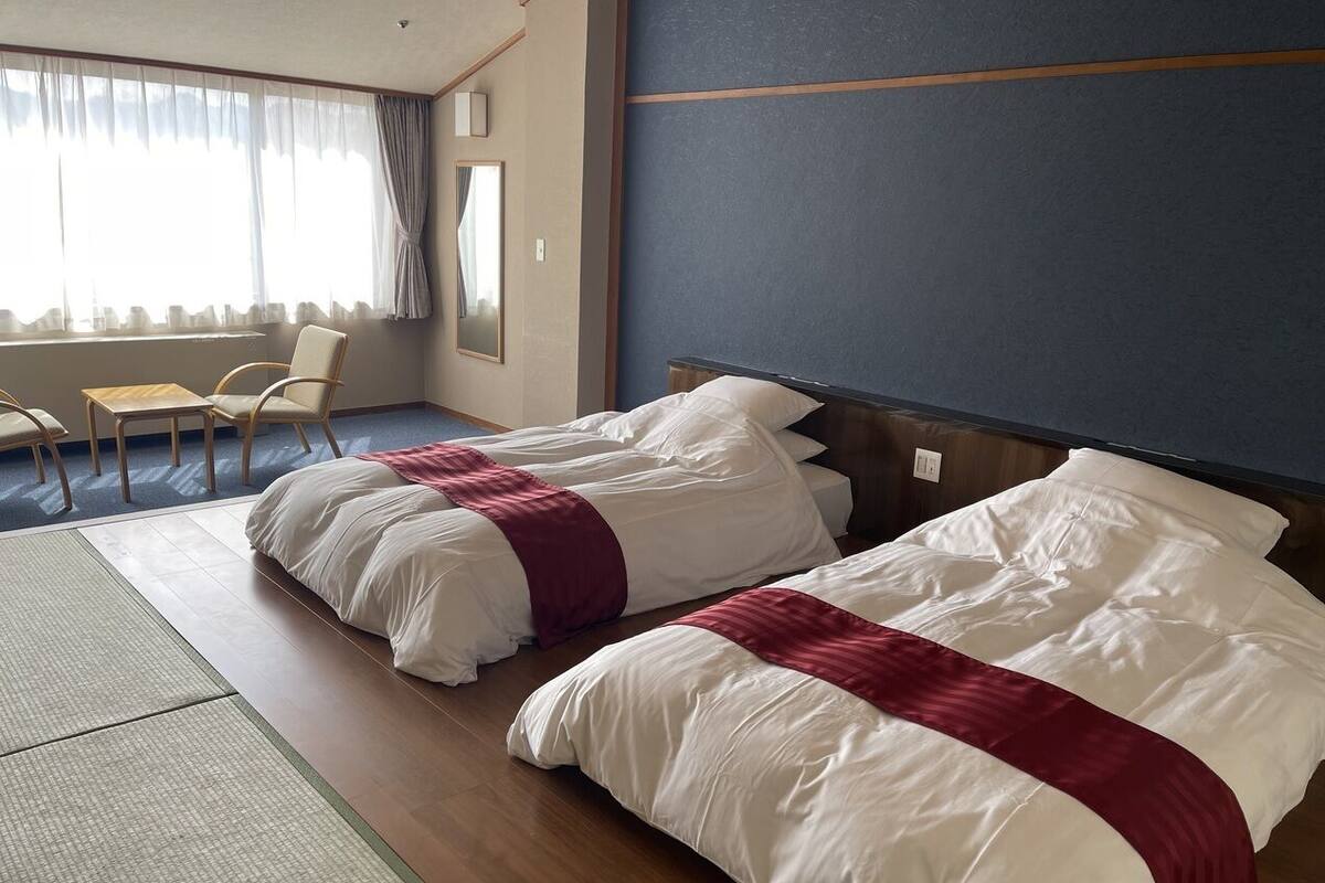 Habitación de hotel con dos camas, zona de estar con sillas, un ventanal grande con cortinas y suelo de madera.