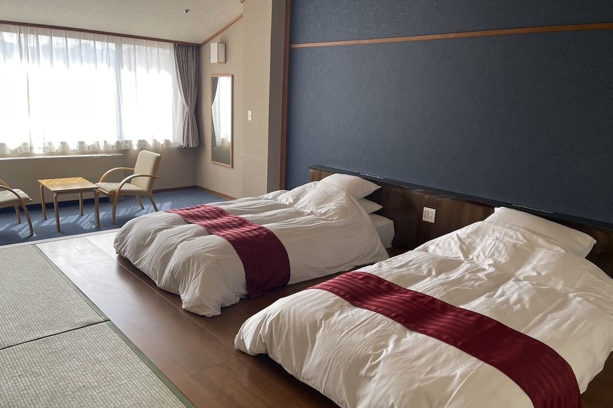 A hotel room with two beds, a sitting area with chairs, a large window with curtains, and a wooden floor.