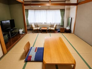 Quarto tradicional em estilo japonês, com tatames, uma mesa de madeira baixa, duas cadeiras, televisão e uma pequena cozinha.