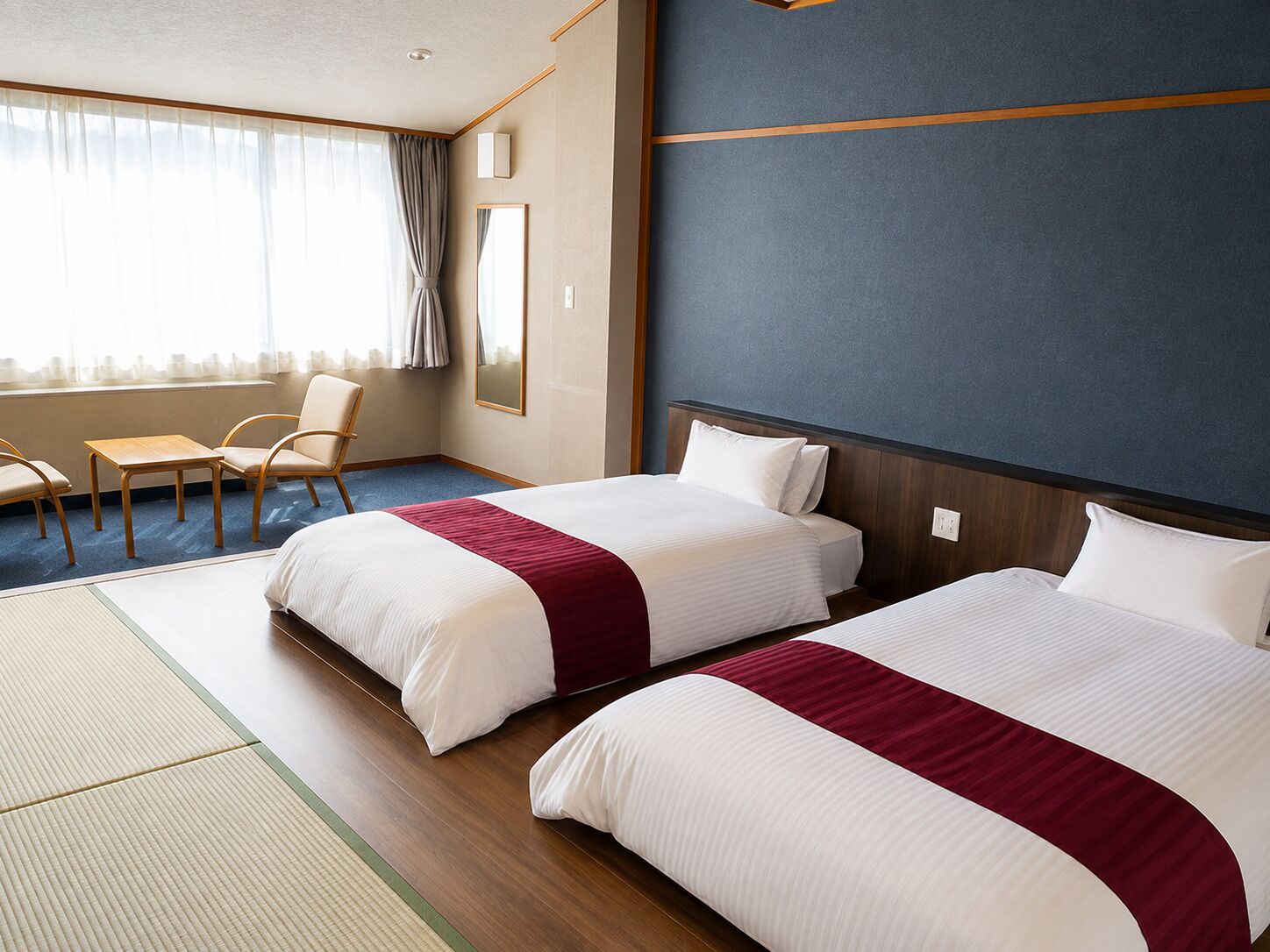 Habitación de hotel con dos camas, un rincón de descanso con una silla y una mesa, y un ventanal con cortinas.