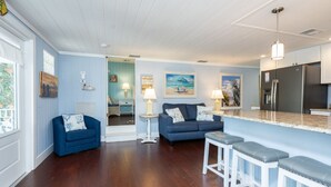 A modern kitchen with a stainless steel refrigerator, a dining area with a blue sofa and a small table, and a bar area with stools. - Beachwalker Apartment:  Duplex within 100 steps to the sand. (Holmes Beach)