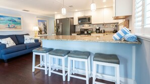 A modern kitchen with white cabinets, stainless steel appliances, and a granite countertop. There is a blue sofa and three white barstools with gray cushions in the living area. - Beachwalker Apartment:  Duplex within 100 steps to the sand. (Holmes Beach)