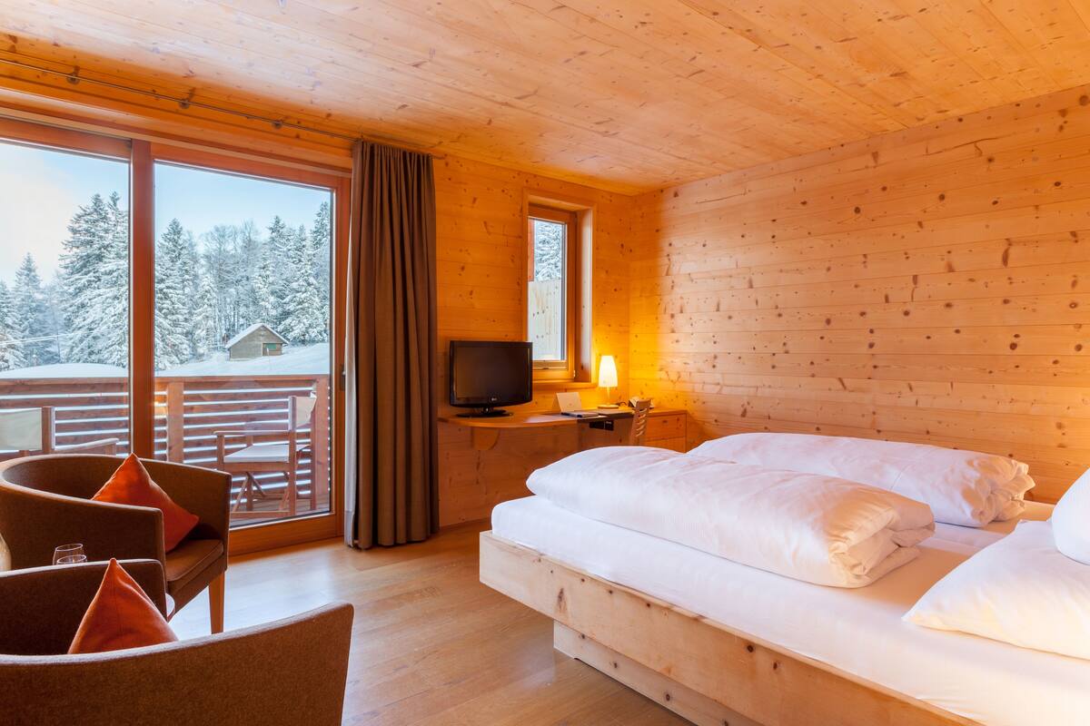 Een houten kamer met een bed, een televisie en een balkon met uitzicht op met sneeuw bedekte bomen.