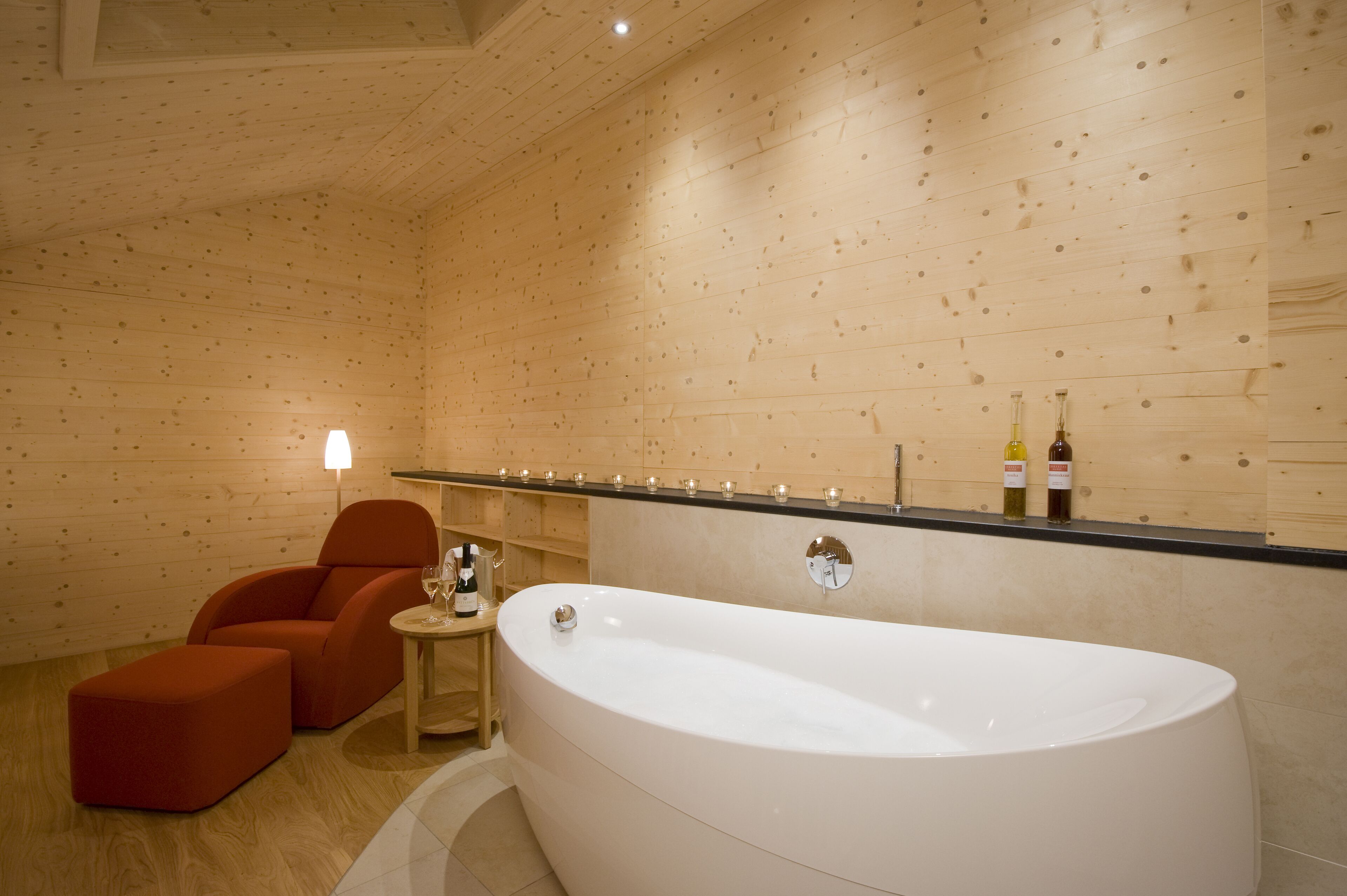 Ein modernes Badezimmer mit Holzvertäfelung, einer großen Badewanne, einem kleinen runden Tisch mit Flasche und Glas sowie zwei roten Stühlen.
