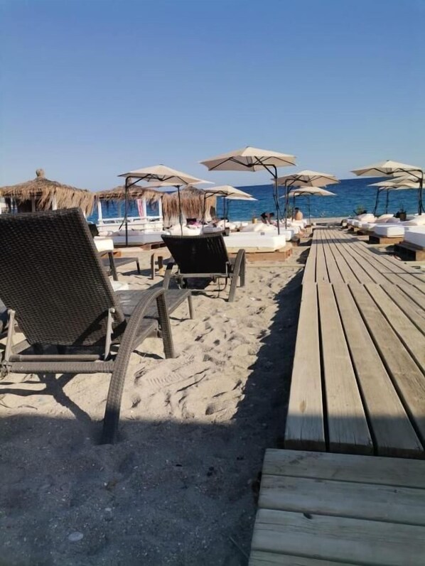 A beachside area with wooden deck chairs and umbrellas, a thatched-roof structure, and a view of the ocean.