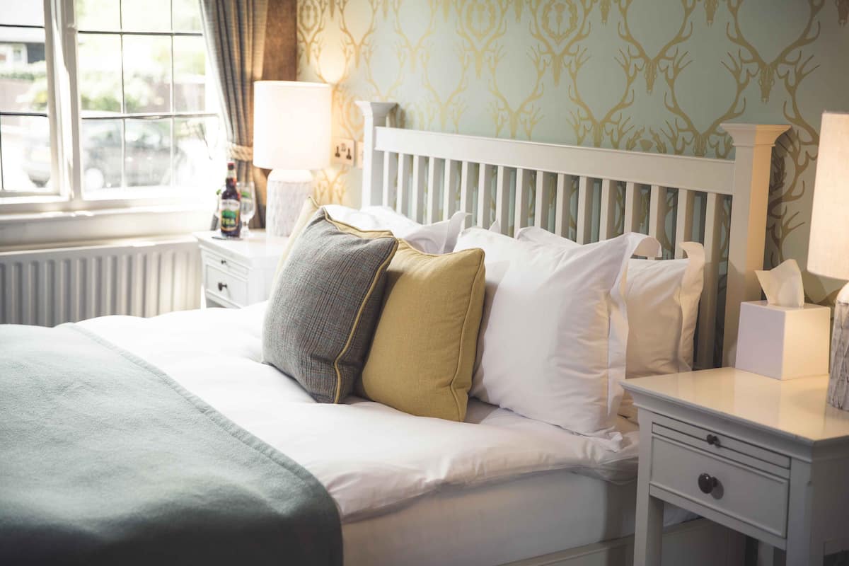 A neatly made bed with white bedding and a variety of pillows, a bedside table with a lamp, and a window with curtains.