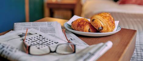 Une table en bois dressée pour le petit-déjeuner, avec des croissants sur une assiette, un journal et une paire de lunettes.