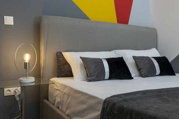 A neatly made bed with a grey headboard, a bedside table with a lamp, and a power outlet.