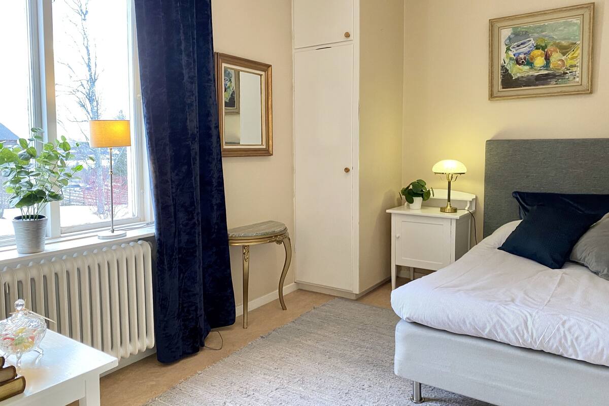 Une chambre à coucher avec un lit, une table de chevet avec une lampe, un petit tabouret, un radiateur, une fenêtre avec des rideaux, un miroir et un tableau accroché au mur.