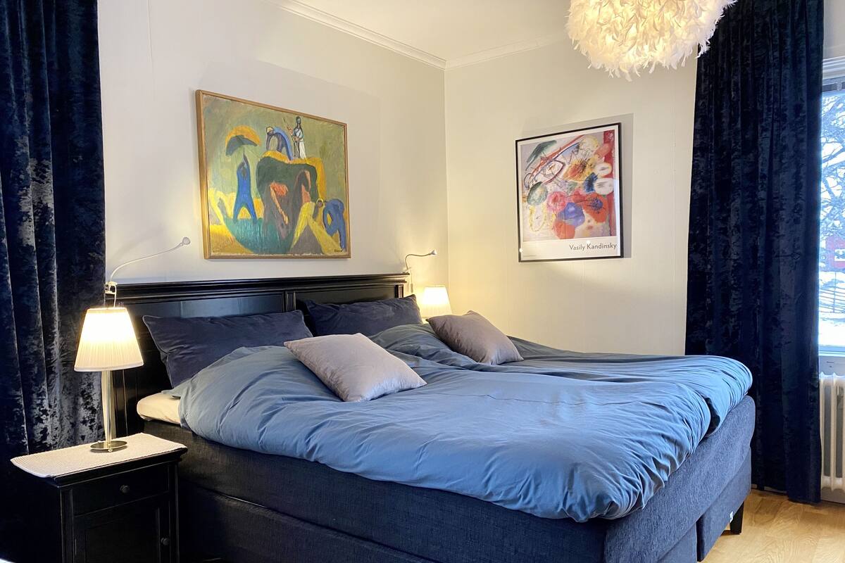Une chambre à coucher avec un grand lit, deux tables de chevet avec des lampes et deux œuvres d’art encadrées accrochées au mur.