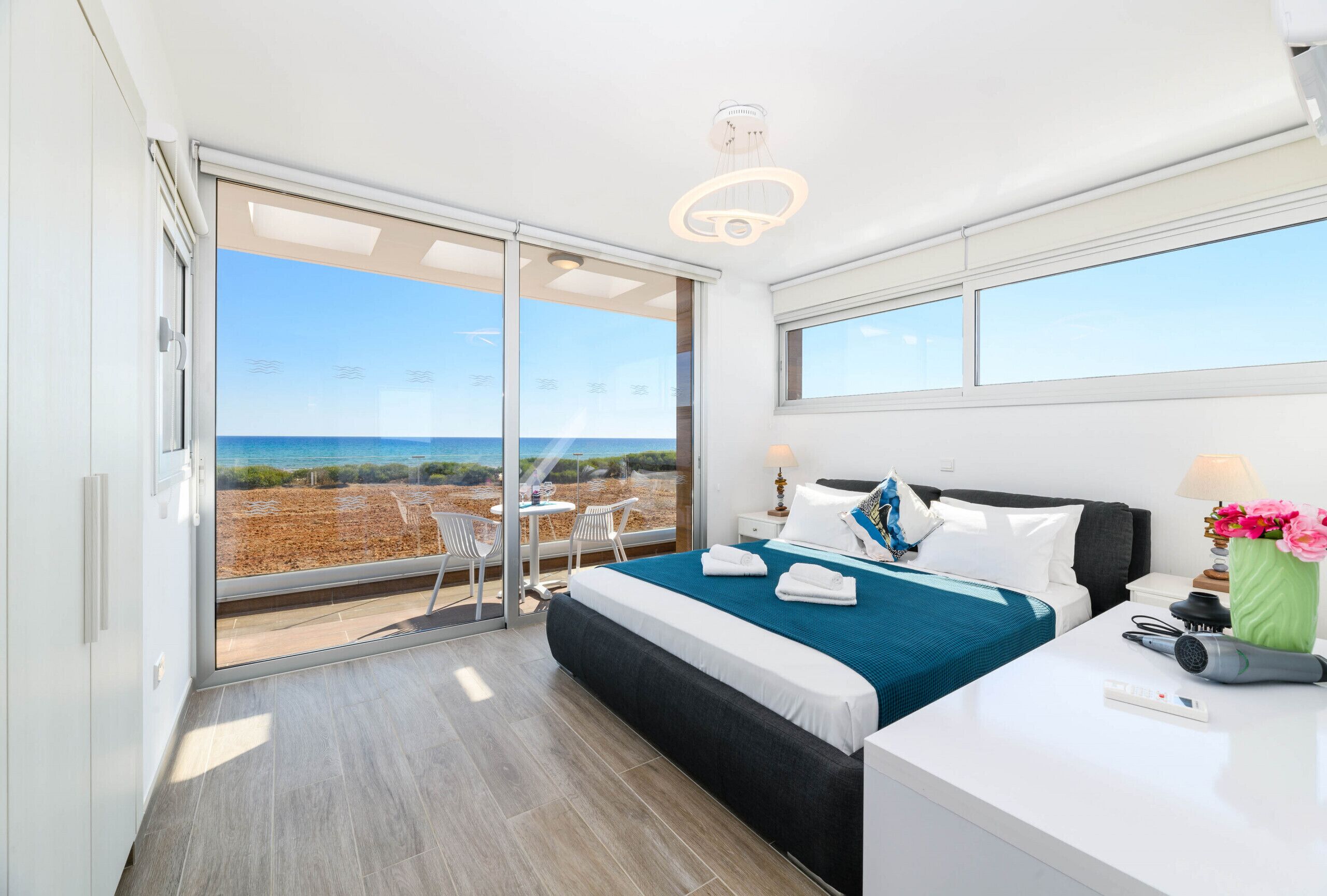Een moderne hotelkamer met een grote schuifdeur naar uitzicht op het strand, een net opgemaakt bed, een klein tafeltje met een lamp en een haardroger op de nachtkastje.