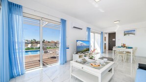 A modern hotel room with a large sliding glass door to a pool area, a flat-screen TV, a dining table with chairs, and a white console table with a bowl and cups. - Infinity View Villa - Charming Ayia Thekla Getaway (Ayia Napa)