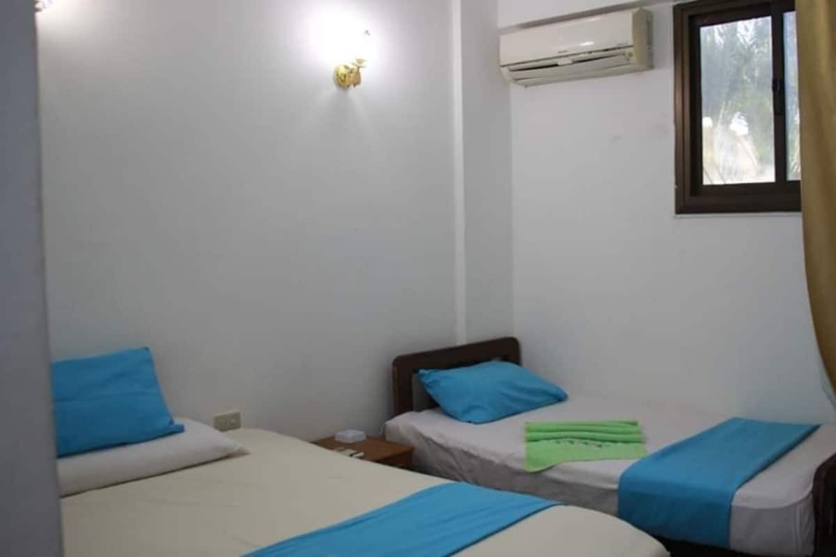 A small, clean room with two single beds, a window with curtains, an air conditioning unit, and a wall-mounted light fixture.