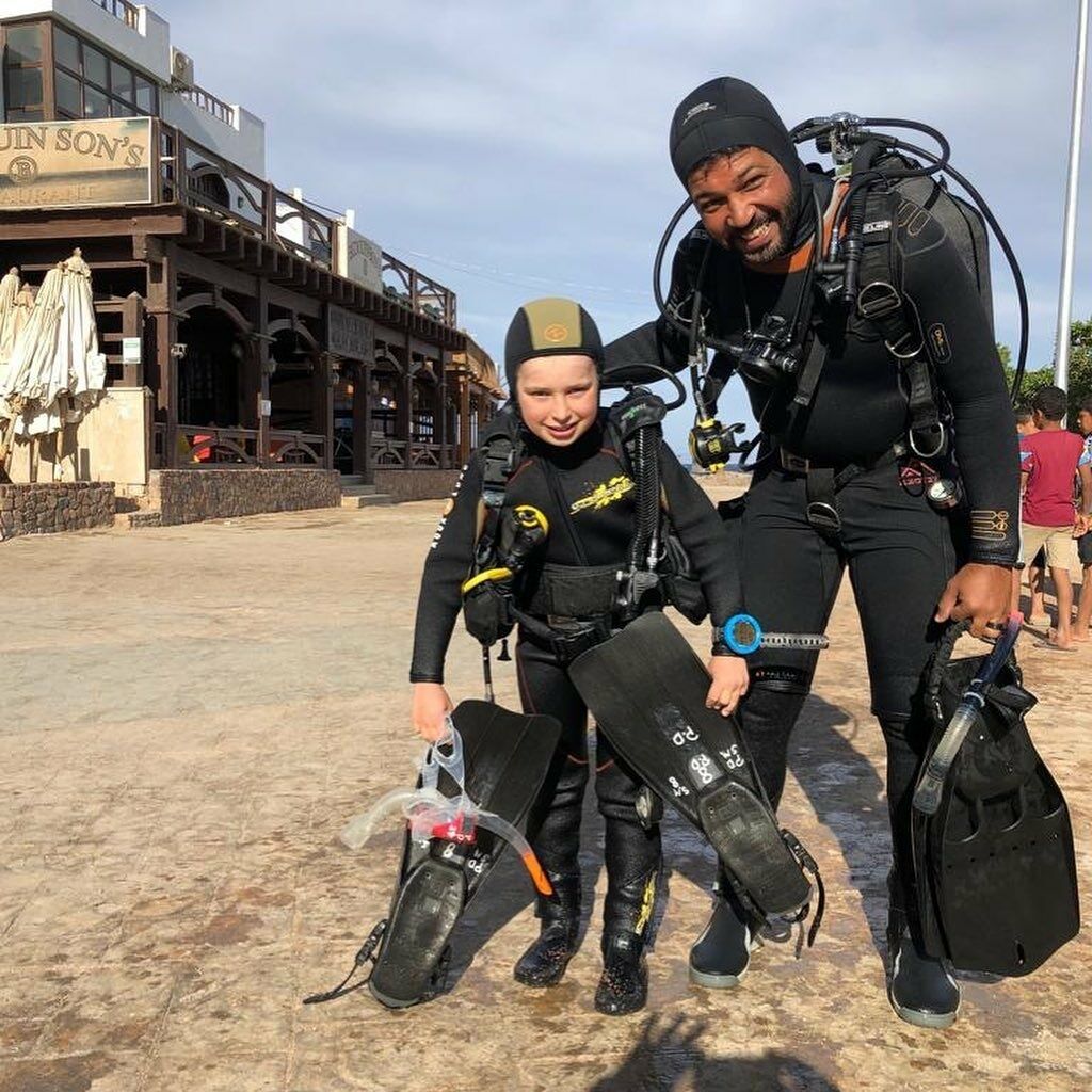 Una persona con traje de neopreno y equipo de buceo de pie junto a un niño con traje de neopreno, ambos sujetando aletas de buceo.