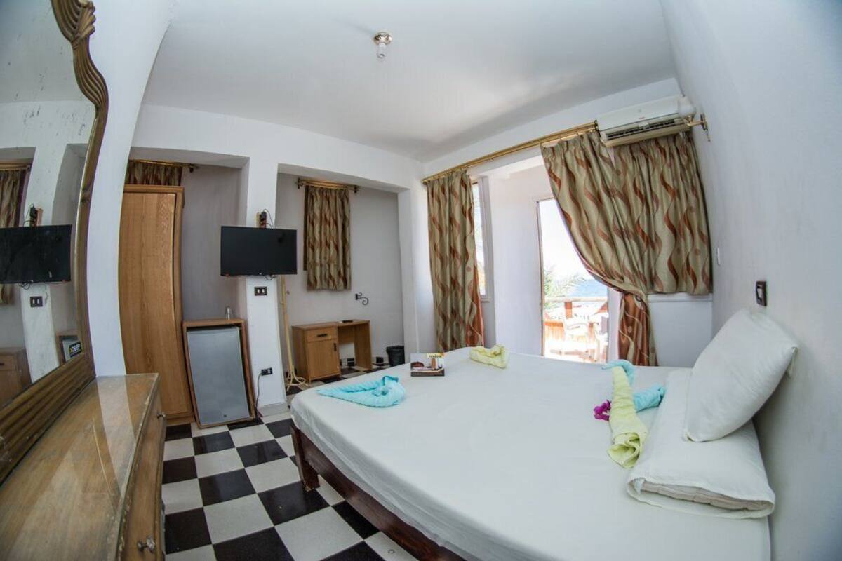 A hotel room with a checkered floor, a bed with pillows, a TV, a desk, and a wardrobe.
