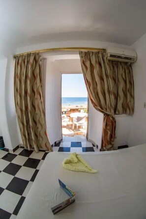 A hotel room with a checkered floor, a bed with a white sheet, a yellow towel, and a view of the beach through a doorway. - Penguin Village Dahab (Dahab)