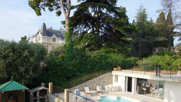 A swimming pool with a stone wall and black railing, surrounded by greenery and a view of a large building in the background. - Comfortable villa - 3 rooms - PRIVATE swimming pool in a quiet location in Nice (NICE)