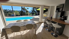 A modern kitchen with a bar area, a poolside dining setup, and a view of a swimming pool and greenery.