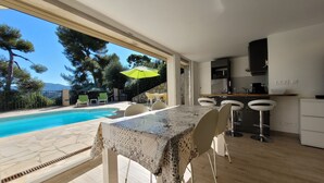 A modern kitchen with a bar area, a dining table with chairs, and a pool area with a view of trees and mountains. - Comfortable villa - 3 rooms - PRIVATE swimming pool in a quiet location in Nice (NICE)