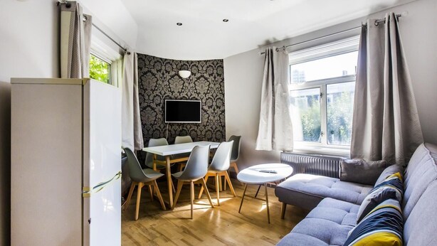 A modern living room with a grey sofa, a dining table with chairs, a TV mounted on the wall, and a window with curtains. - My City Chic St. Pancras Apartment (London)