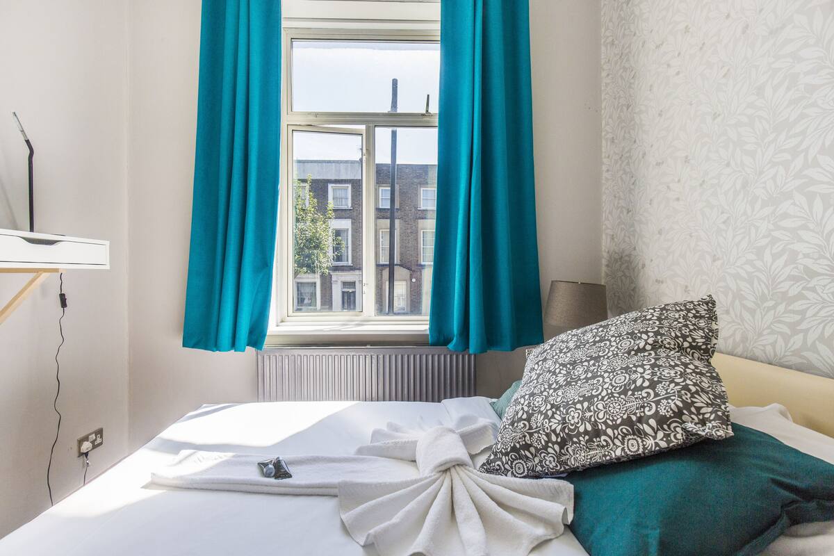 Un lit bien fait, agrémenté d’un coussin à motifs, une fenêtre avec des rideaux de couleur turquoise et un radiateur.
