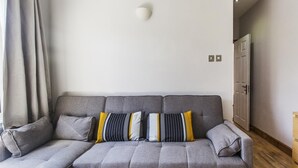 A grey sectional sofa with multiple cushions in a neutral-toned room with a wooden floor and a white door. - My City Chic St. Pancras Apartment (London)