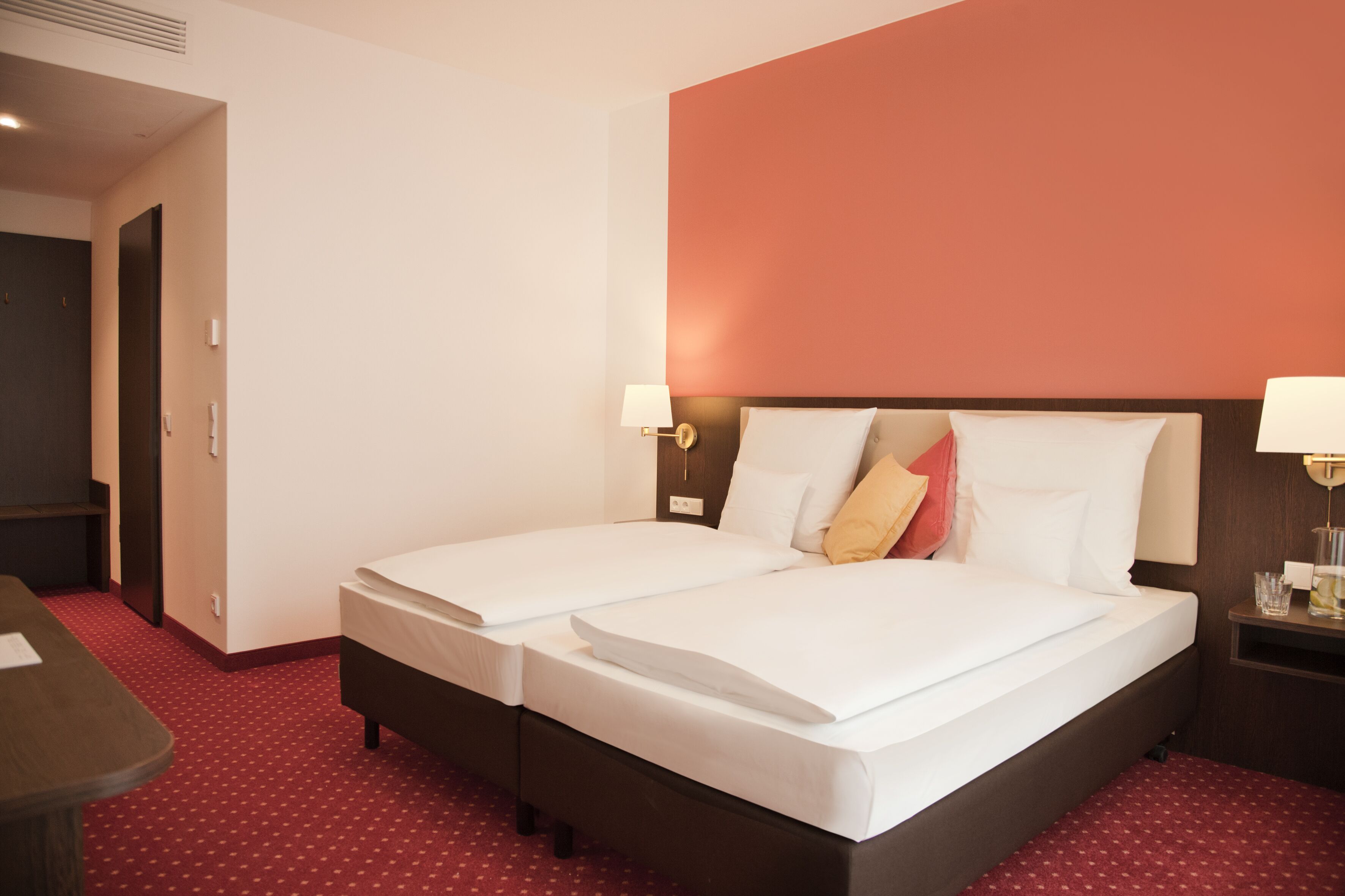 Habitación de hotel con dos camas, una pared de acento roja y una mesita de noche pequeña con lámpara.