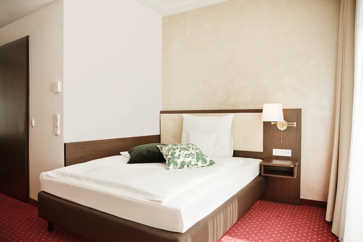 Hotellihuone, jossa on suuri sänky, yöpöytä, lamppu ja punaisella matolla päällystetty lattia.