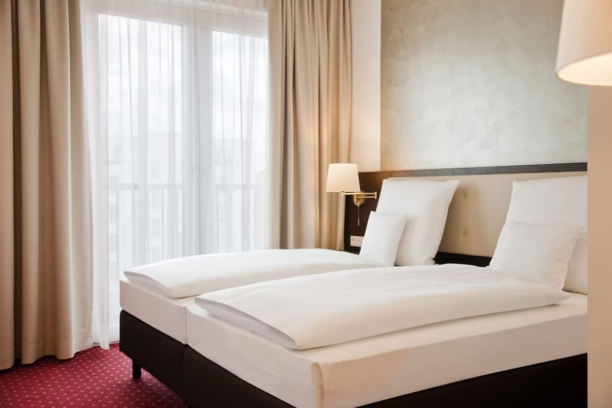 Hotellihuone, jossa on parivuode, yöpöytälamppu, ikkuna verhoilla ja punainen matto.