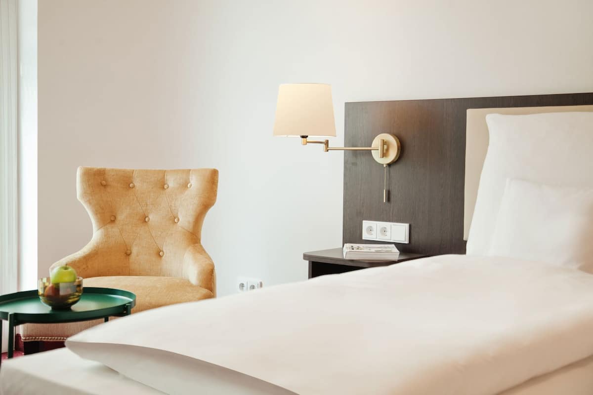 Hotellihuone, jossa on sänky, tuoli, yöpöytä lampun kanssa ja seinälle asennettu puhelin.
