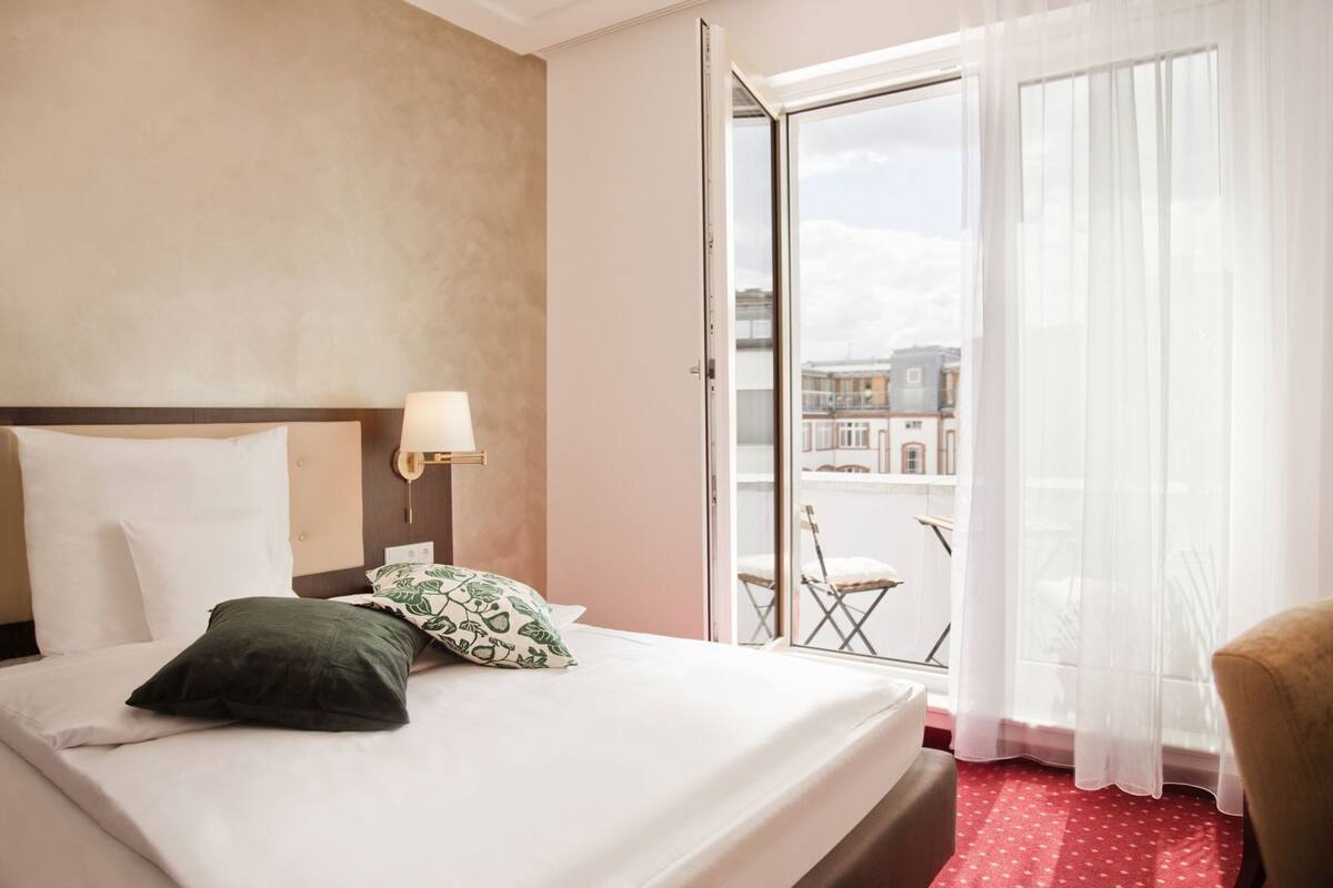 Hotellihuone, jossa on sänky, yöpöytä, lamppu ja parvekkeelta avautuva näkymä kaupunkiin.