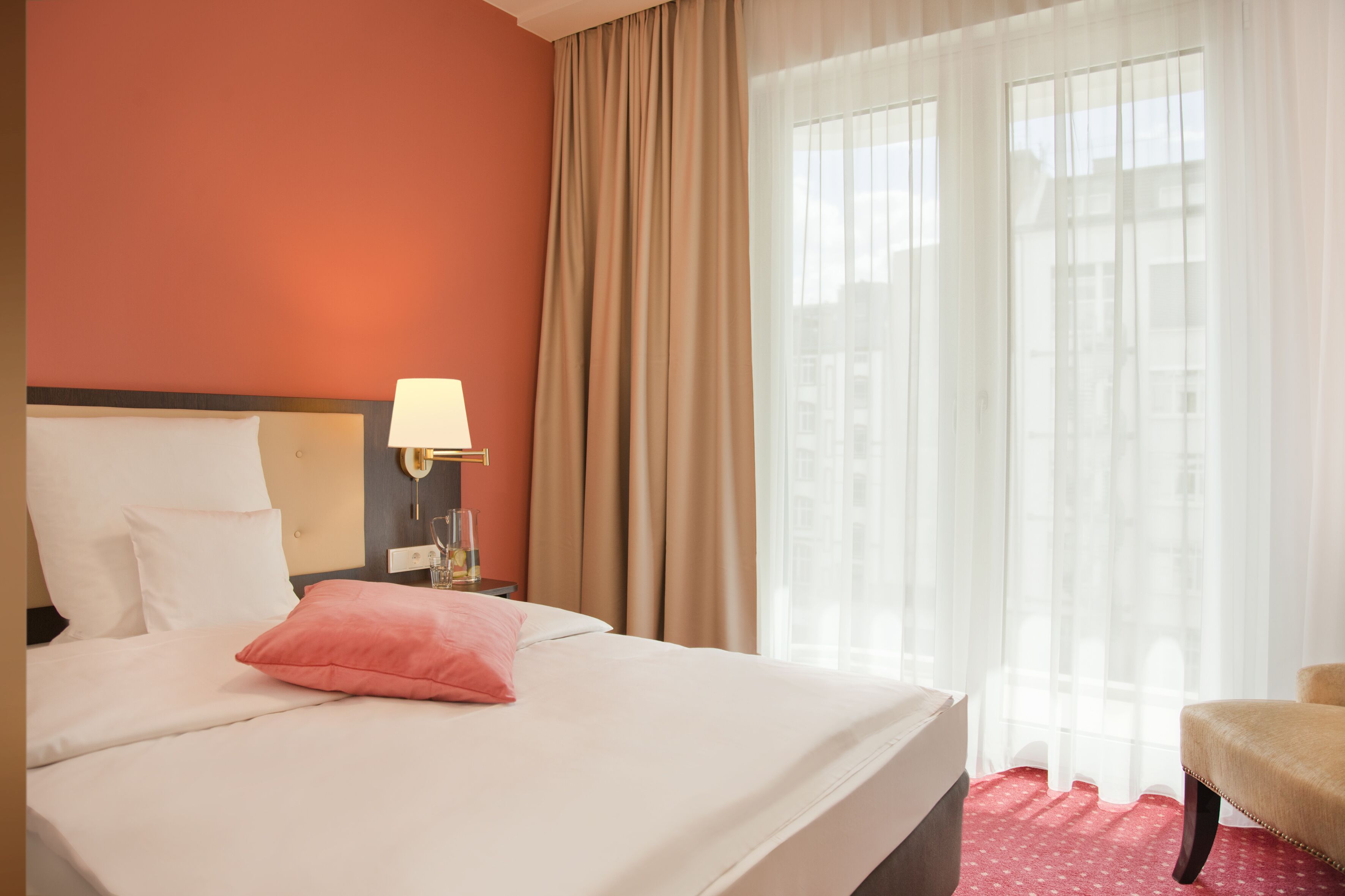 Una habitación de hotel con cama, mesita de noche con lámpara, una silla y una ventana con cortinas.