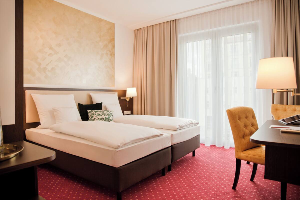 Hotellihuone, jossa on kaksi sänkyä, työpöytä, tuoli ja suuri ikkuna, jossa on verhot.