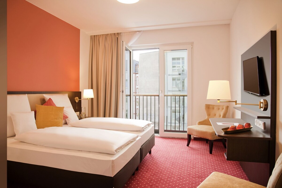 A hotel room with a bed, a desk with a TV, a chair, and a balcony with curtains.