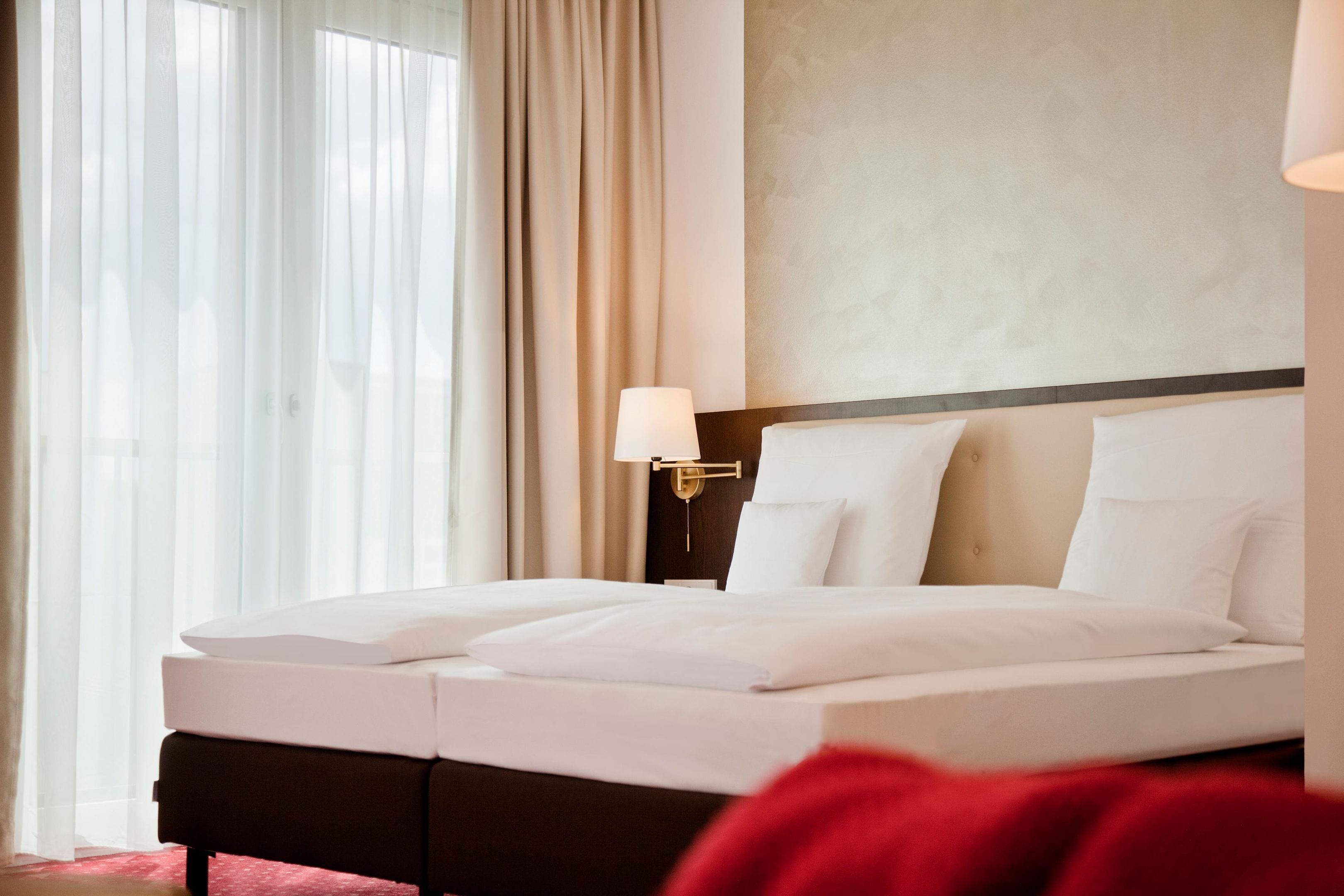 Una habitación de hotel con una cama bien hecha, una lámpara de noche y un ventanal con cortinas.