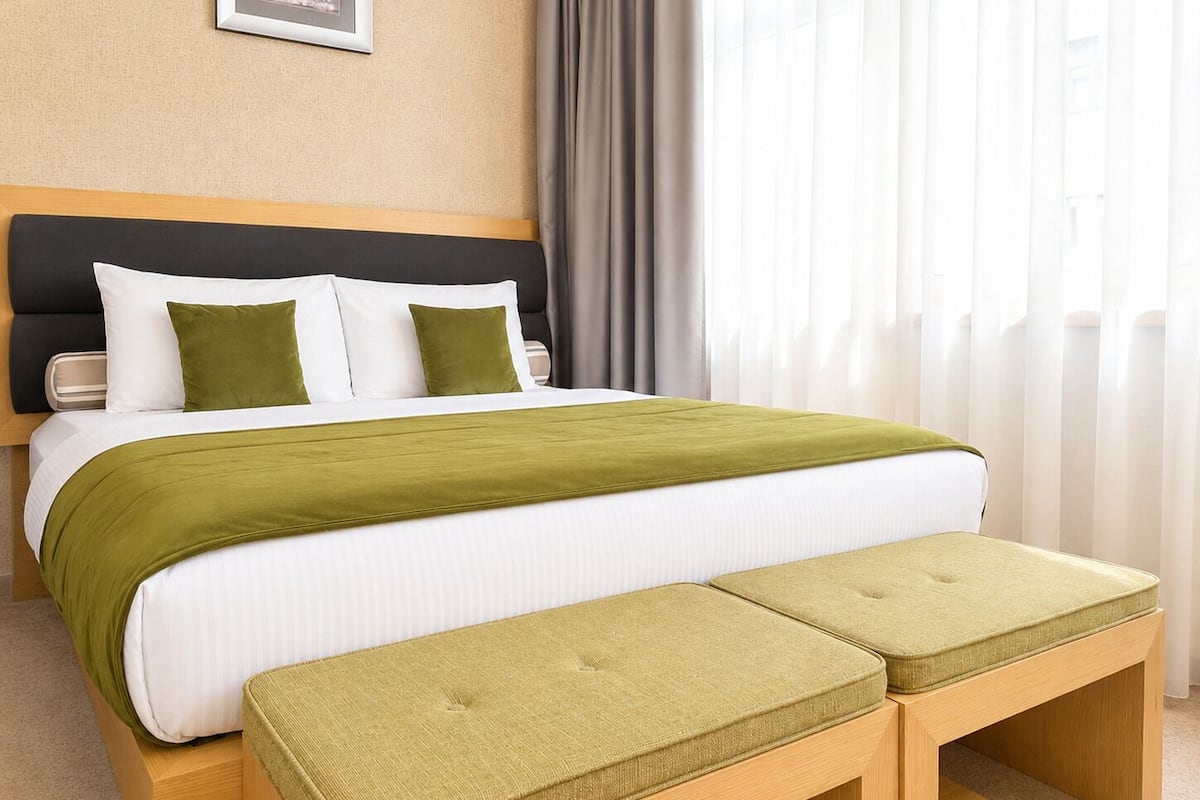 A hotel room with a bed, a headboard, a bedside table, a bench, and a window with curtains.