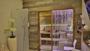 A modern spa room with a wooden floor, a massage table, a white chair, and a glass partition with a tree design. - Atera Business Suites (Belgrade)