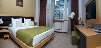 Habitación de hotel con cama, mesitas de noche, escritorio, silla, ventana con cortinas y vistas a edificios.