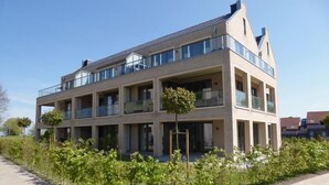Un immeuble résidentiel à plusieurs étages, doté de balcons, d’une allée pavée et de jeunes arbres au premier plan.