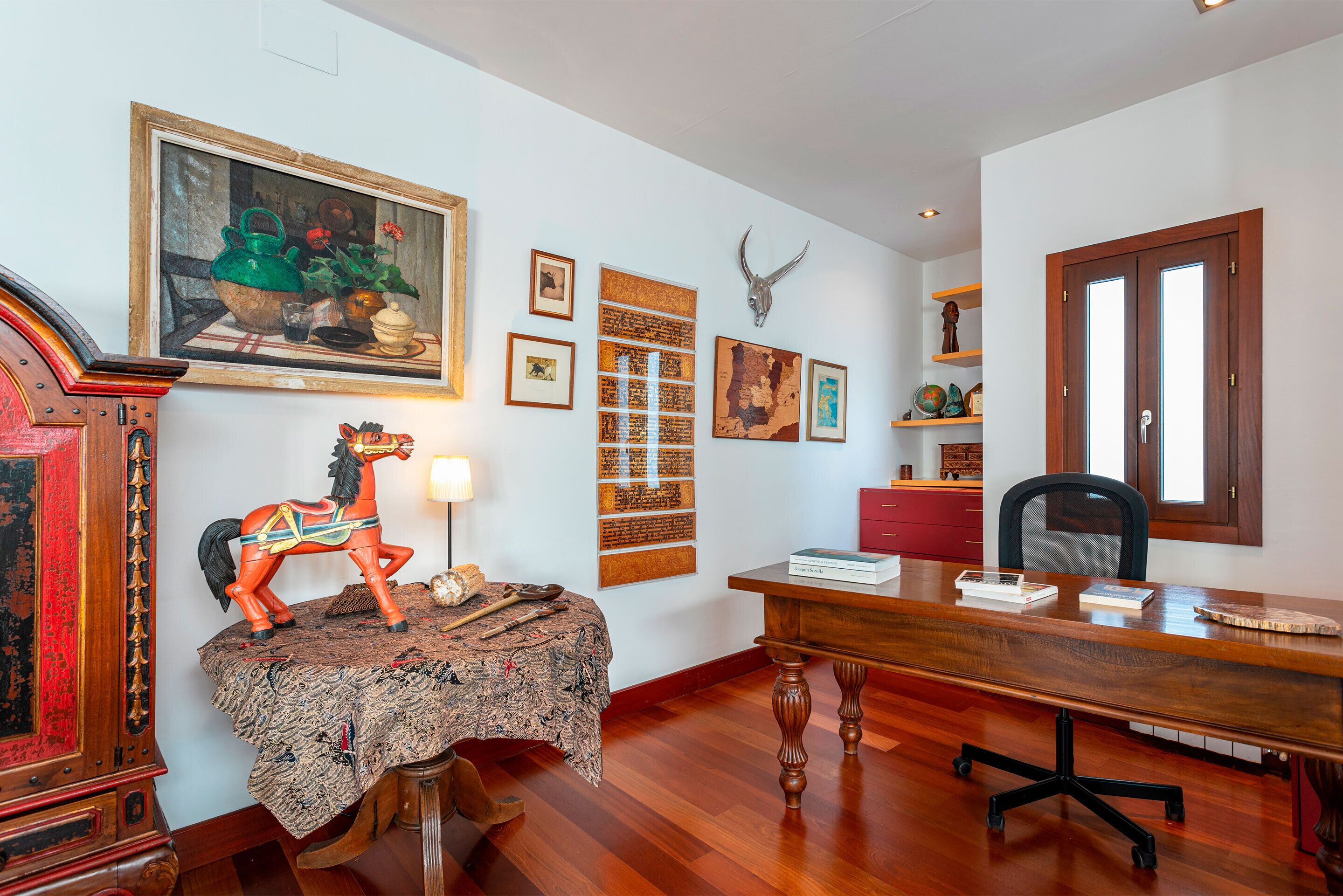Una habitación con un escritorio de madera, una silla, una mesita con una figurita de caballo y varias imágenes enmarcadas en la pared.