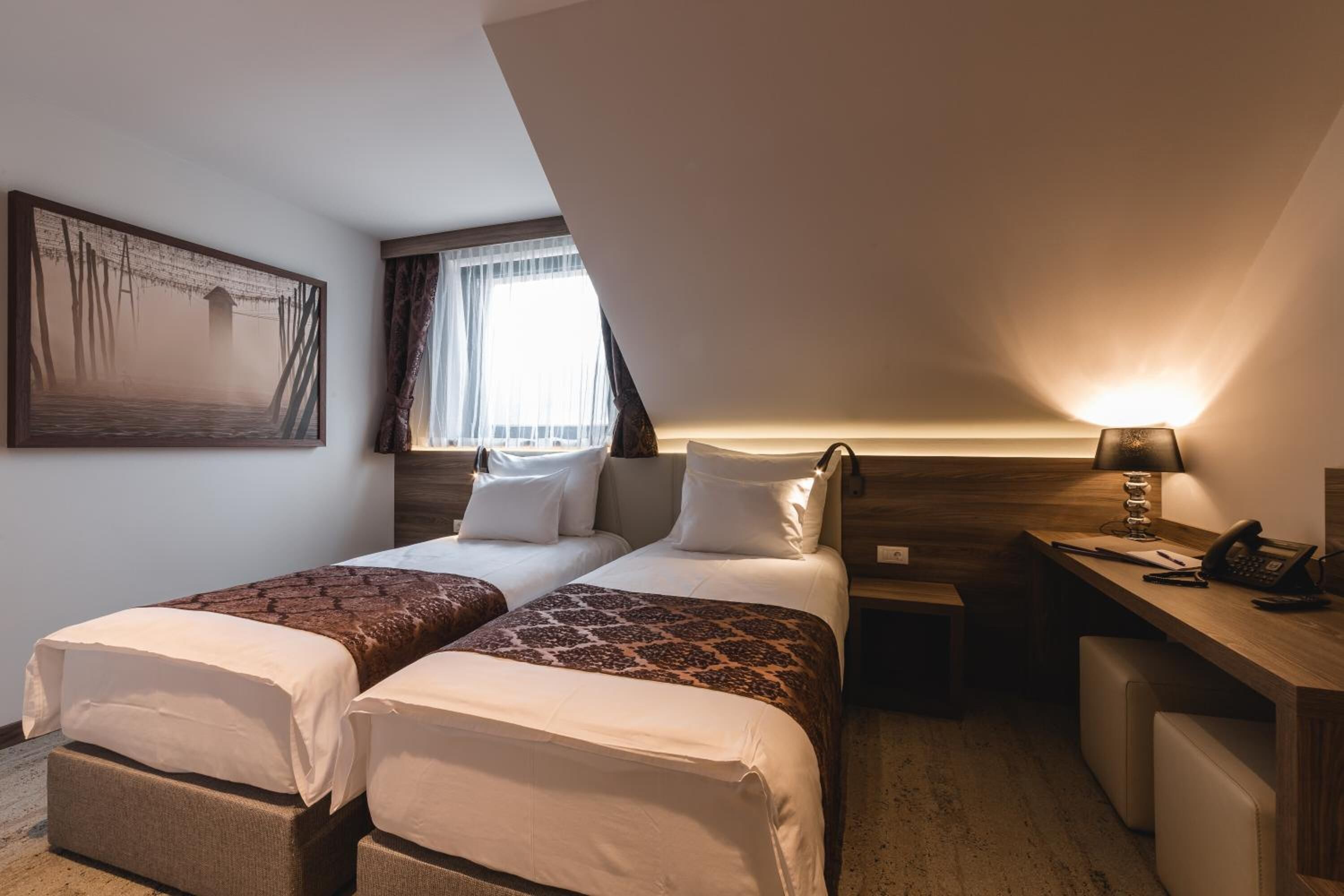 Quarto de hotel com duas camas, uma mesa, uma luminária e um quadro na parede.