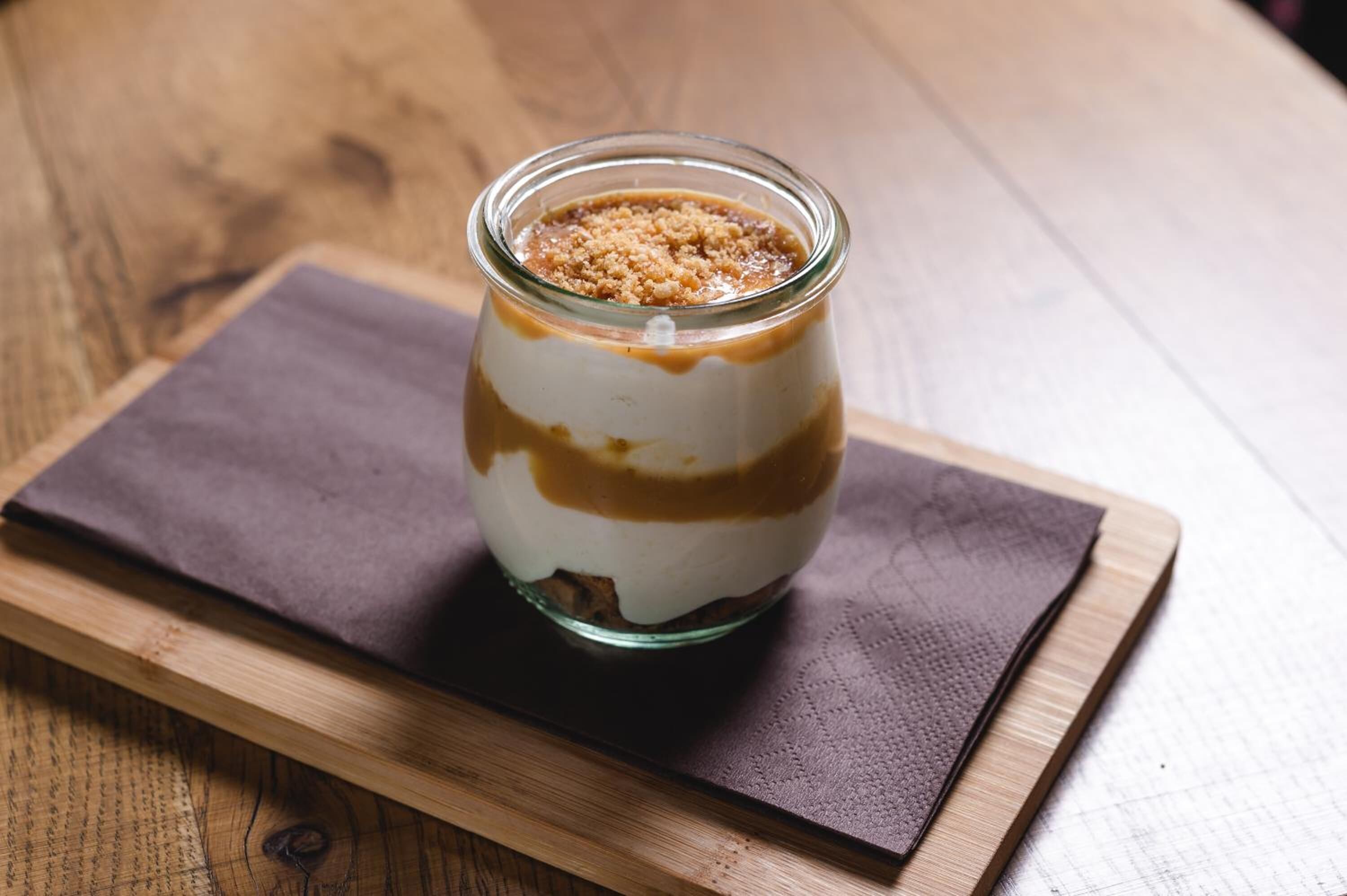 Postre en un tarro de vidrio con cobertura marrón, sobre una tabla de madera con tela oscura debajo.