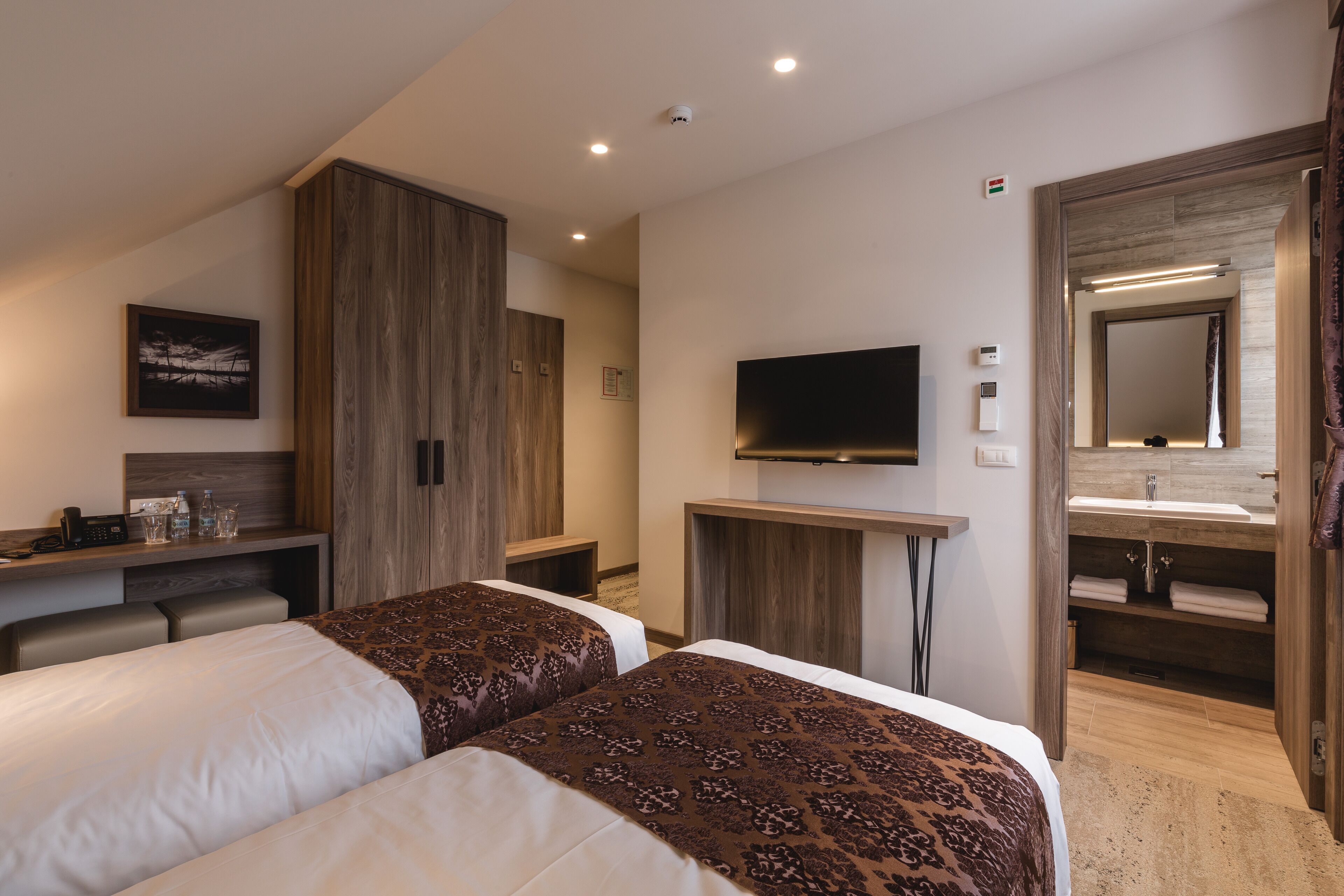 A hotel room with two beds, a desk with a flat-screen TV, a wooden wardrobe, a bathroom with a mirror and sink, and a small seating area.