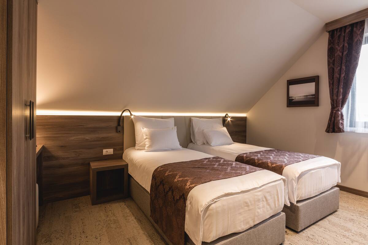 A bedroom with two beds, a nightstand, a bedside table, a wall-mounted lamp, a framed picture, and a window with curtains.