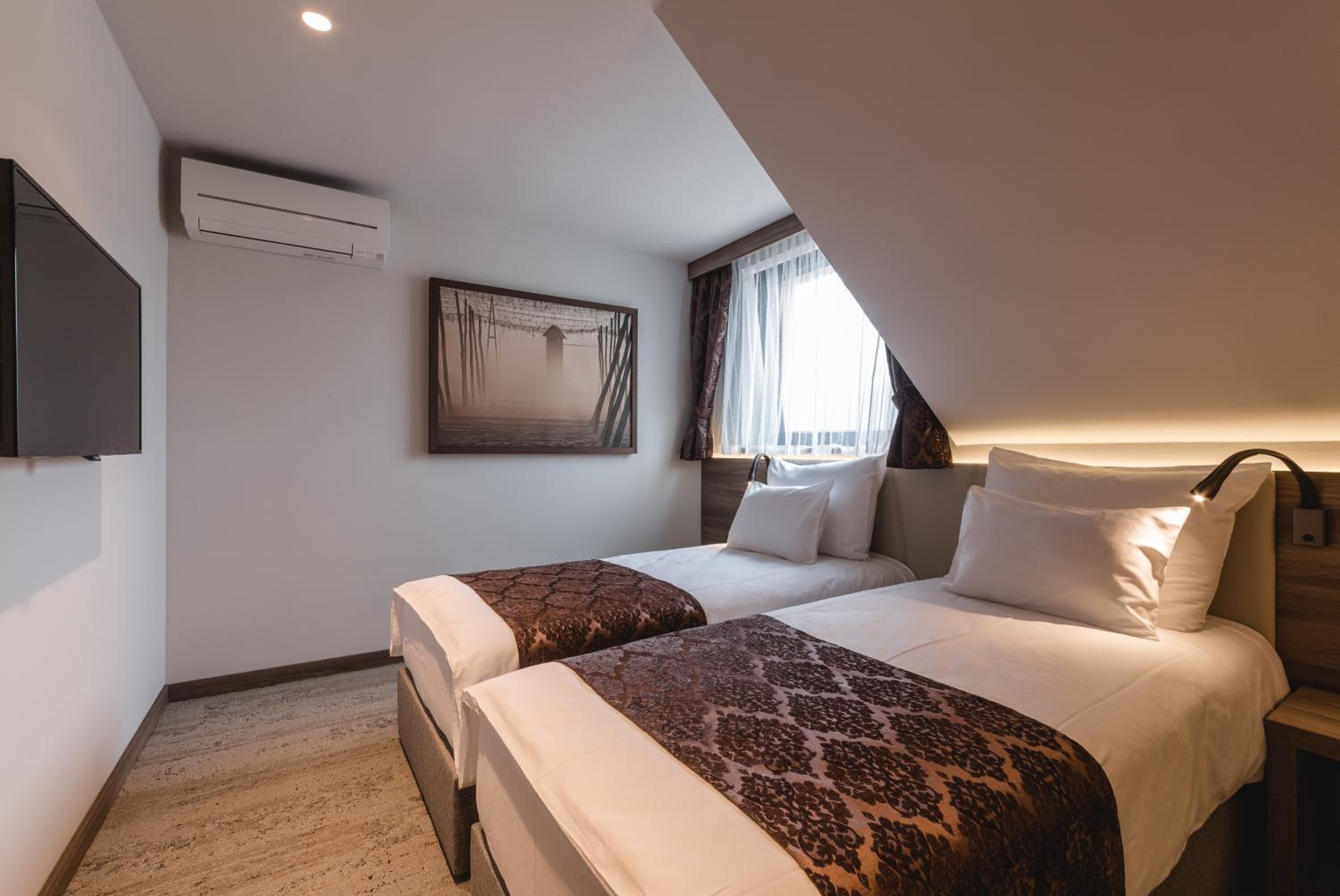 A hotel room with two beds, a mounted TV, an air conditioning unit, and a painting on the wall.