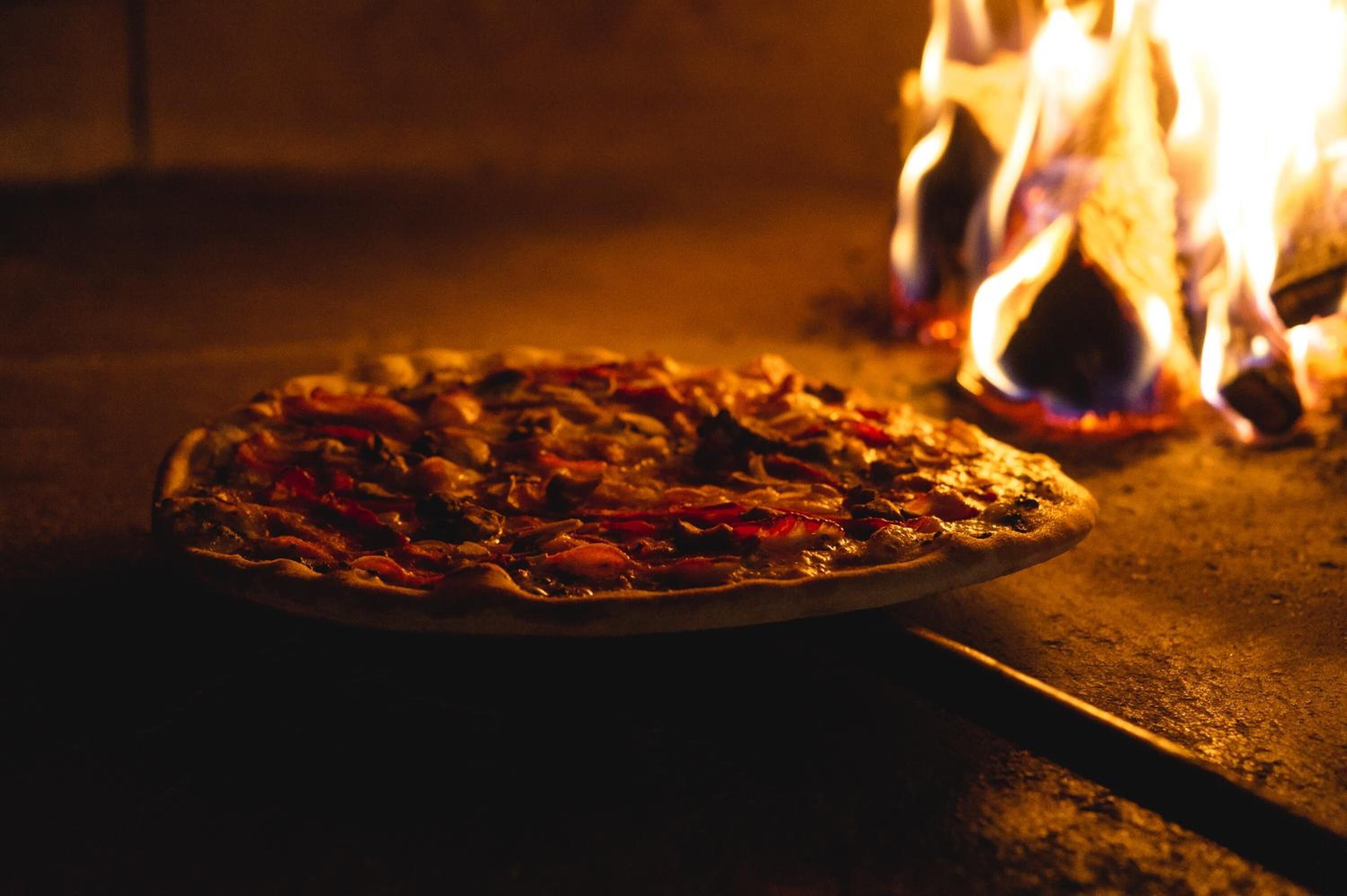 A pizza in a wood-fired oven with flames visible in the background.