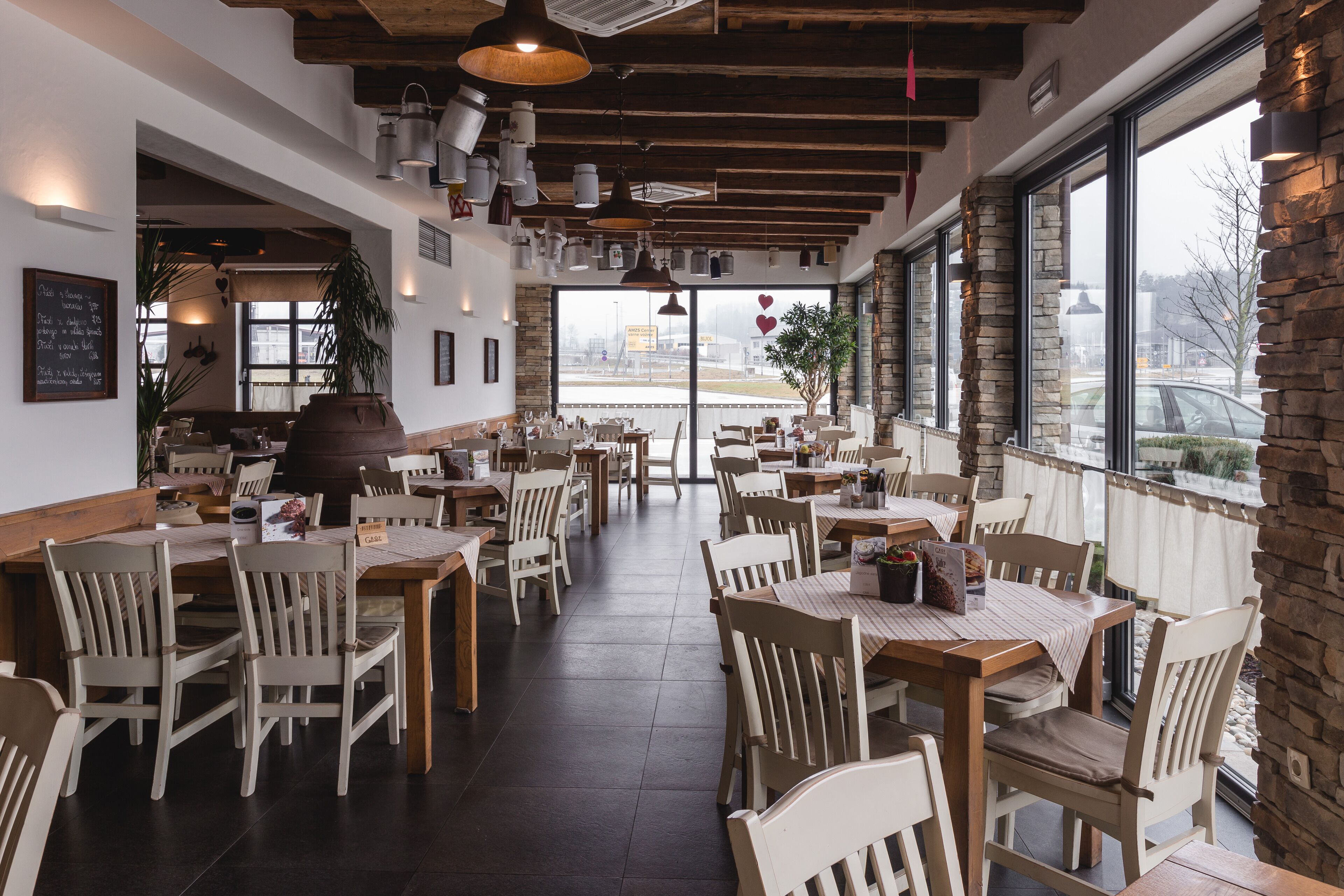 Un restaurante con mesas y sillas de madera, ventanas amplias y una pared de acento de piedra.