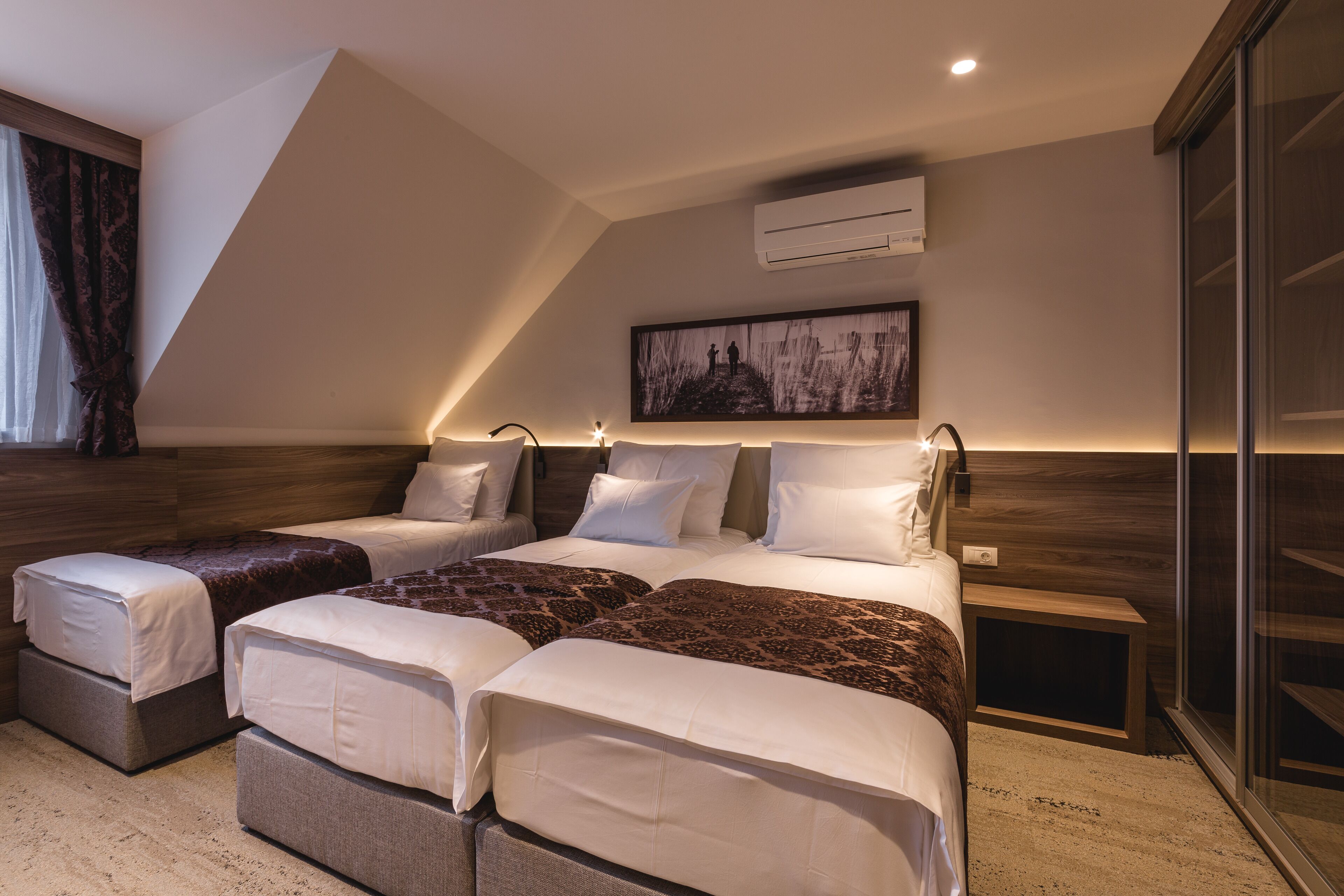 A modern bedroom with a sloped ceiling, a large bed with multiple pillows, a bedside table, and a wall-mounted air conditioner.