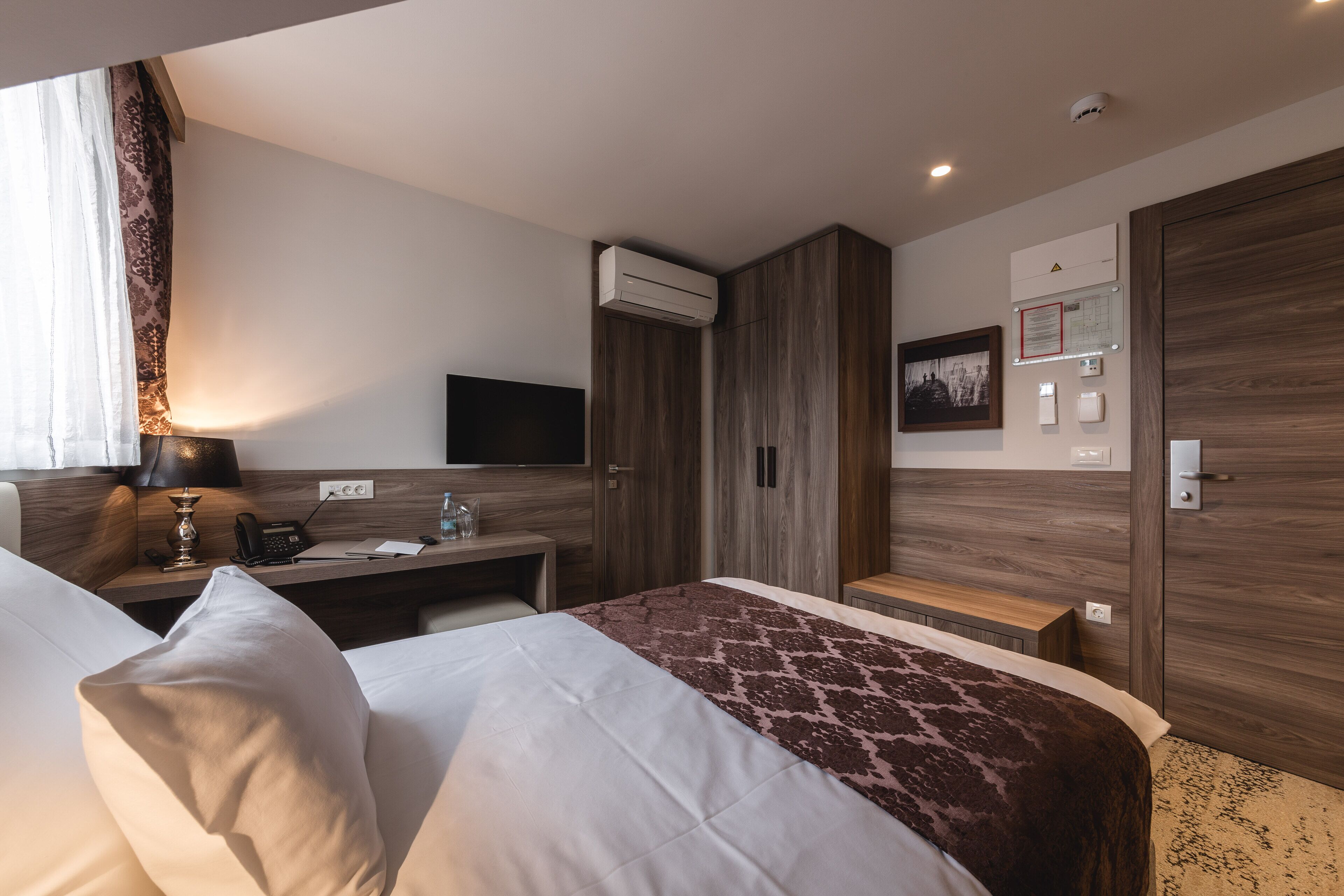 A hotel room with a bed, bedside table, lamp, air conditioning unit, and a wall-mounted TV.