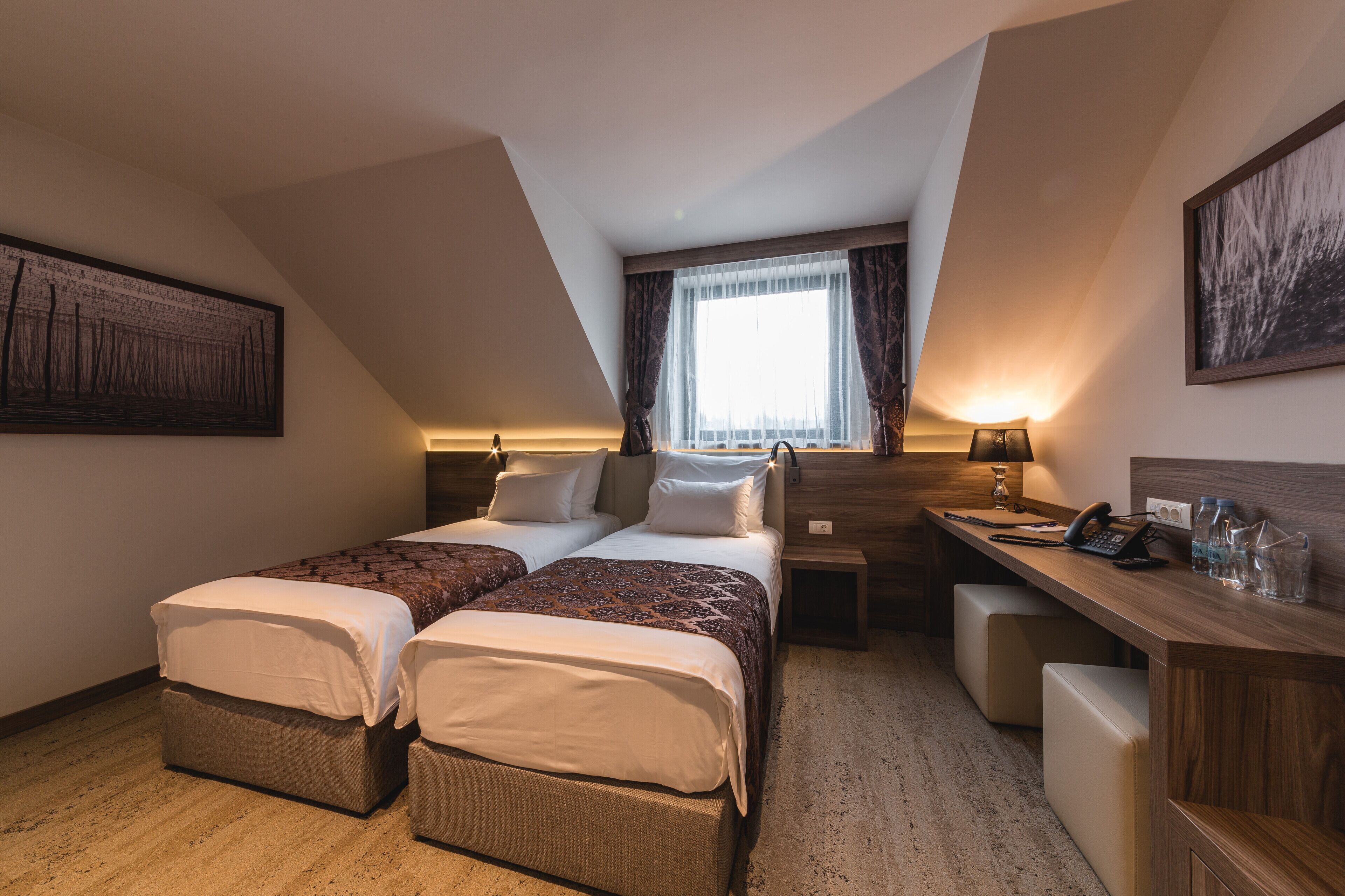 Une chambre d’hôtel avec deux lits, un bureau, une chaise, une lampe et une fenêtre avec des rideaux.